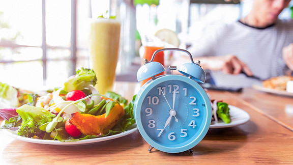 Intermittierendes Fasten ist gemäß dem aktuellen Stand der Forschung eine sinnvolle Methode, die möglicherweise zu einem längeren Leben beitragen kann. Symbolbild für intermittierendes Fasten mit einem Smoothie und Salat und einem dekorativen Wecker.